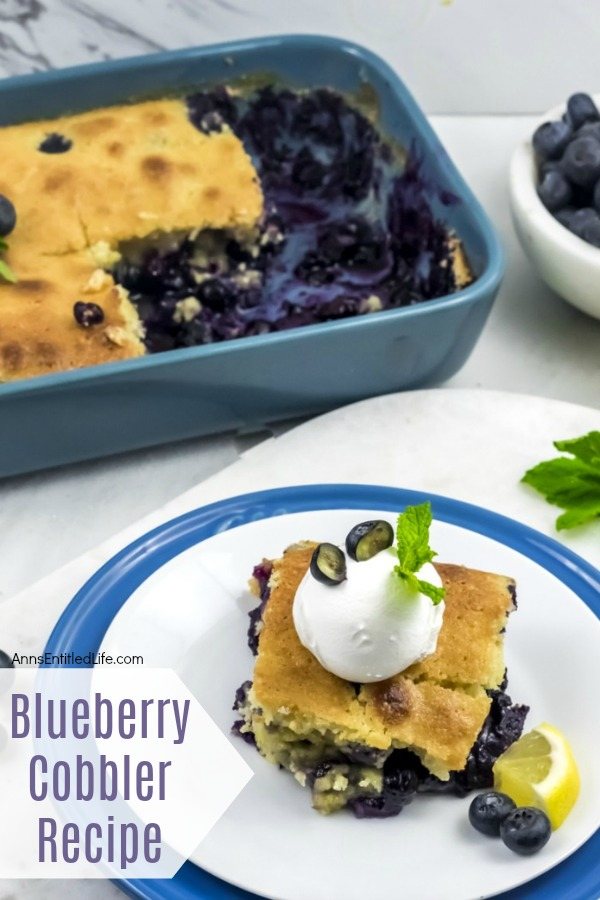 Overhead view of a blue plate underneath a white plate, a serving of blueberry cobbler on top of the white plate. The remaining pan of blueberry cobbler is in the upper left, a white bowl of fresh blueberries is in the upper right.