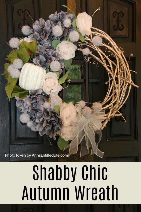 A beautiful fall wreath in colors of grey, blush, and white on a grapevine wreath, hanging on gold wreath hanger against a black door with a glass insert