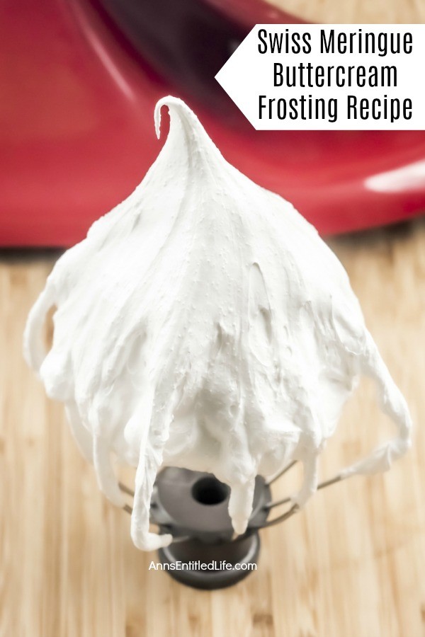 A close-up of a standing whisk beater is in the front and center with white Swiss meringue buttercream frosting adhered to it. There is a red stand mixer in the background. This is on a wooden board.