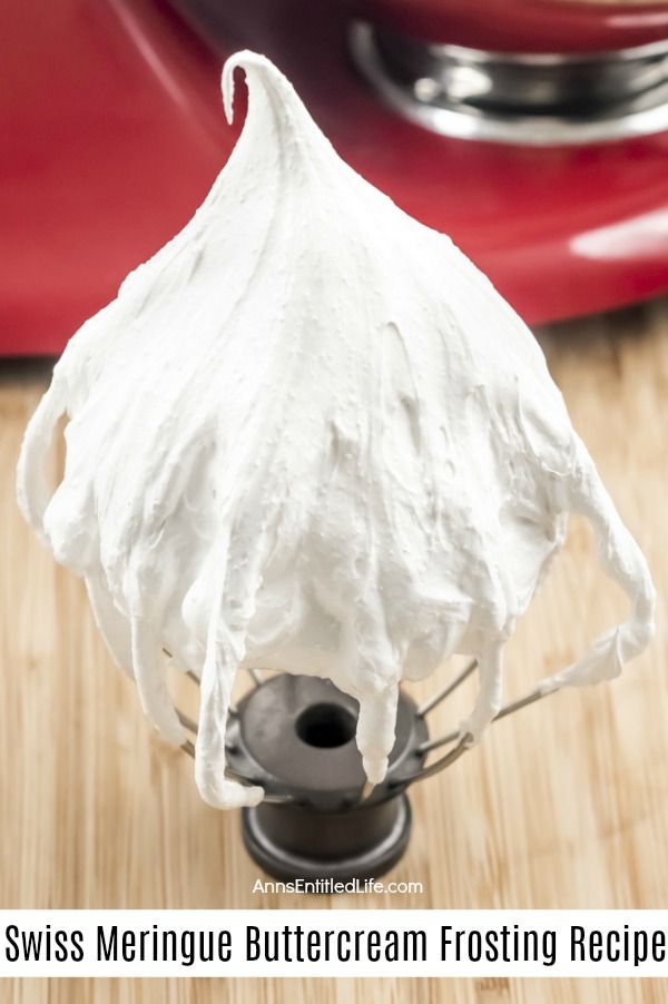 A close-up of a standing whisk beater is in the front and center with white Swiss meringue buttercream frosting adhered to it. There is a red stand mixer in the background. This is on a wooden board.