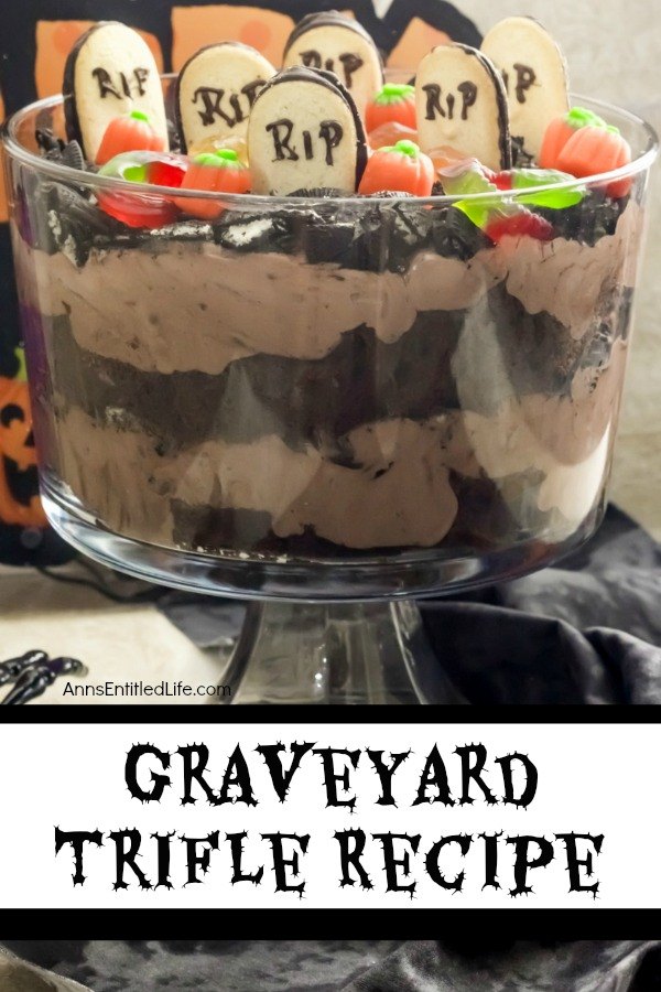 A front-facing view of a layered chocolate trifle with a graveyard setting at the top (grave markers, pumpkins, gummy spiders, and crushed Oreos) in a clear trifle bowl, on top of a black and white table covering. A bone hand is on the left of the trifle bowl.