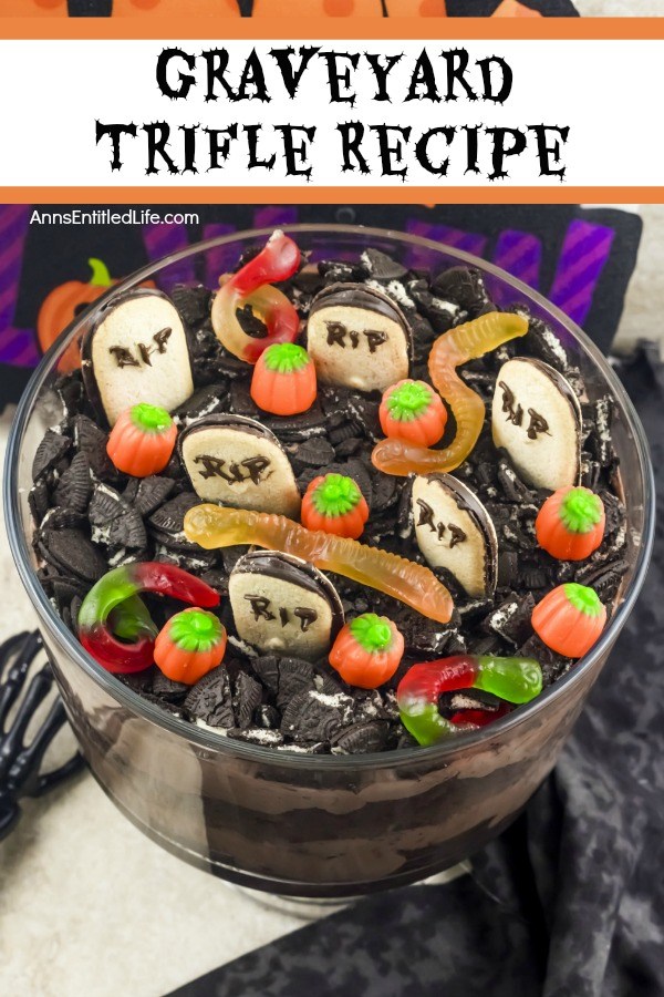 An overhead view of a layered chocolate trifle with a graveyard setting at the top (grave markers, pumpkins, gummy spiders, and crushed Oreos) in a clear trifle bowl, on top of a black and white table covering. A bone hand is on the left of the trifle bowl.