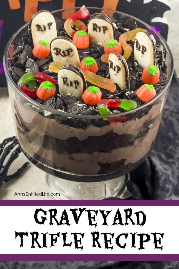 A layered chocolate trifle with a graveyard setting at the top (grave markers, pumpkins, gummy spiders, and crushed Oreos) in a clear trifle bowl, on top of a black and white table covering. A bone hand is on the left of the trifle bowl.