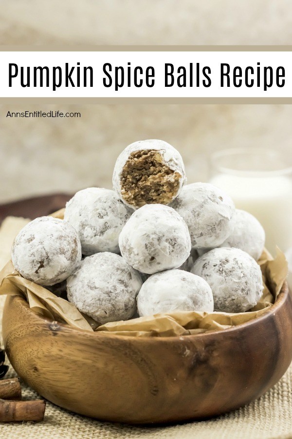 A wooden bowl filled with pumpkin spice balls is center, the top piece has a bite removed, several pieces of cinnamon stick are to the bottom left, a glass of milk in the upper right. All set on a tan placemat, against a brownish background