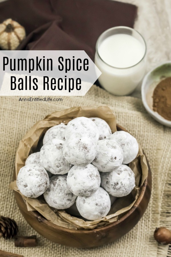 An overhead view of a wooden bowl filled with pumpkin spice balls is center, several pieces of cinnamon stick and a pinecone are to the bottom left, an acorn to the lower right, a glass of milk in the upper right with a small white bowl filled with spice. All set on a tan placemat, against a brownish background