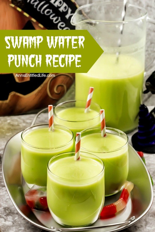 4 glass of green swamp water punch (with an inserted straw in each glass) on a galvanized tray. The remaining swamp water punch is in a pitcher to the right. A Halloween jack-o'-lantern sign is in the back. A few gummy worms are scattered about the tray.