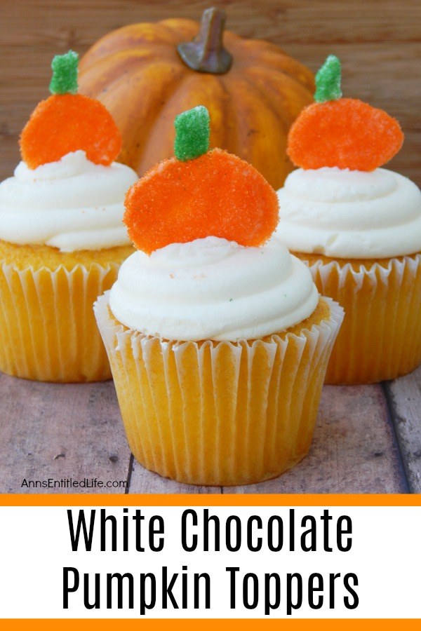 Three white cupcakes with white frosting and a pumpkin topper stuck in the frosting are set on a blue board. There is a faux pumpkin in the background.