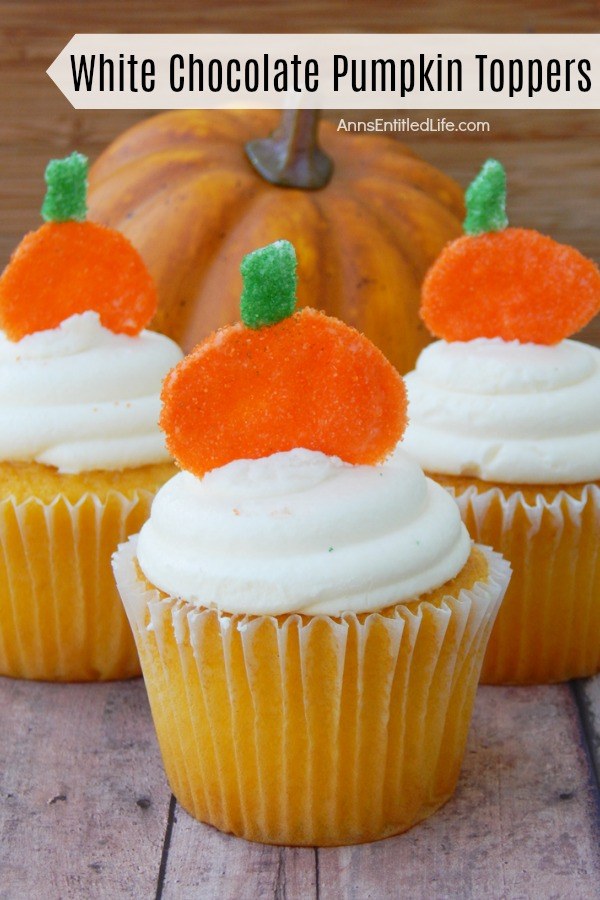 Close-up of three white cupcakes with white frosting and a pumpkin topper stuck in the frosting are set on a blue board. There is a faux pumpkin in the background.