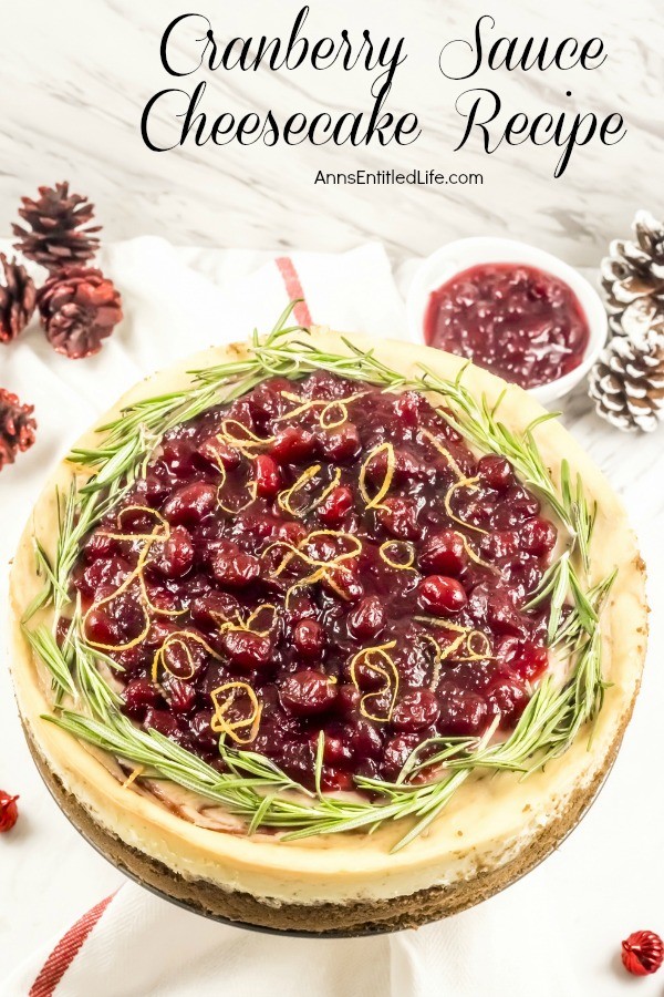 An overhead view of a whole cranberry sauce cheesecake. There are decorative pinecones in the upper right and left, a small red ornament in the lower right, all set against a white background.
