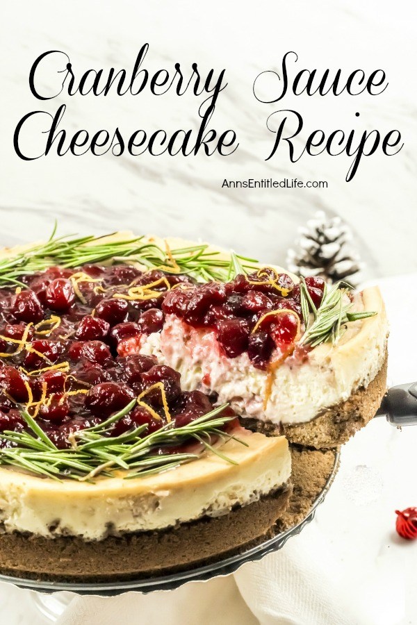 A serving of cranberry sauce topped cheesecake is being lifted from the whole cheesecake with a pie server. There is a pinecone in the upper left, a small red ornament in the lower right.