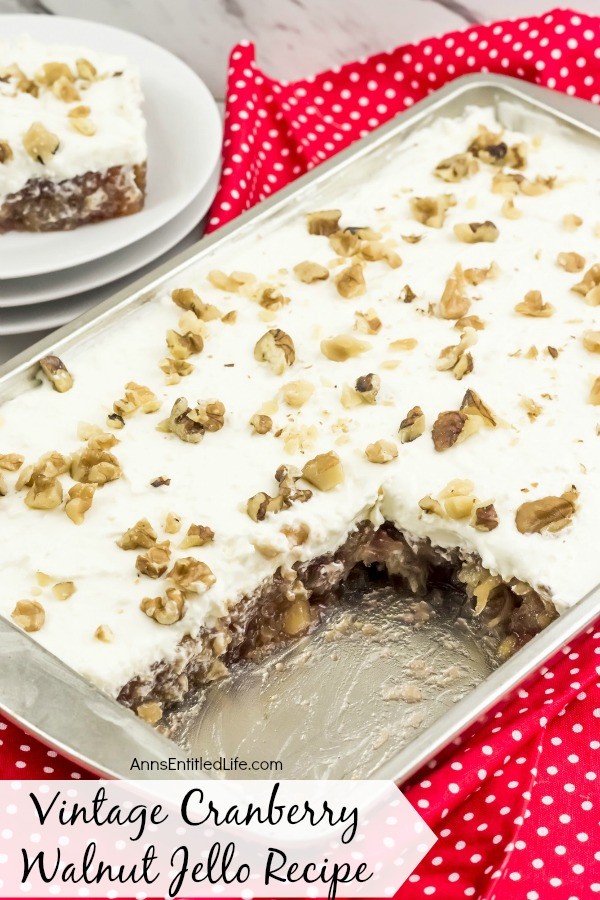 A pan of cranberry walnut jello with a serving missing. That serving is plated in the background, upper left on top of a stack of white dishes. This all sets on a red polka dot napkin.