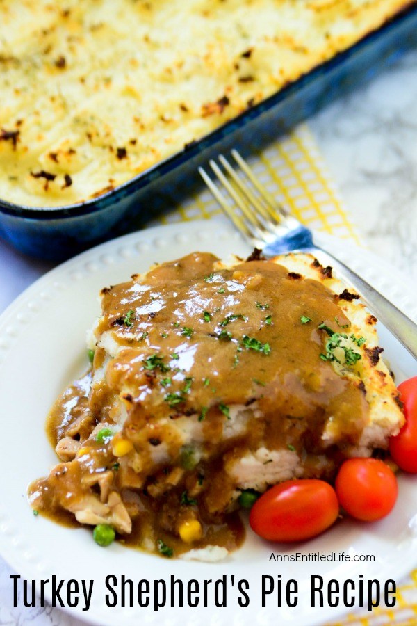 A serving of turkey Shepard pie covered in gravy on a white plate with a fork diagonally across the top of the plate, several tomatoes are garnish at the bottom of the plate. The remaining pie is in a pan in the upper left. A yellow checked dishtowel rests underneath the baking pan.