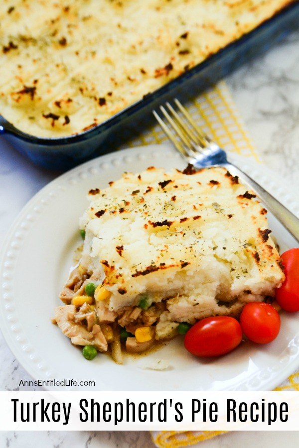 A serving of turkey Shepard pie on a white plate with a fork diagonally across the top of the plate, several tomatoes are garnish at the bottom of the plate. The remaining pie is in a pan in the upper left. A yellow checked dishtowel rests underneath the baking pan.