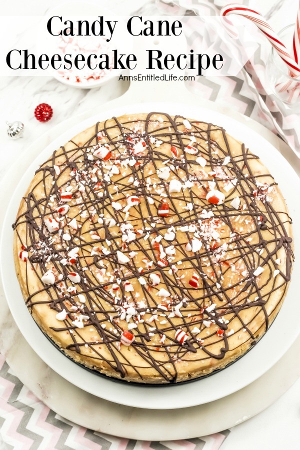 A whole cane candy cheesecake sits atop of white serving platter, sitting on top of a grey chevron-patterned napkin. There are crushed candy candies in a white bowl above the cheesecake, and two whole candy canes to the left.