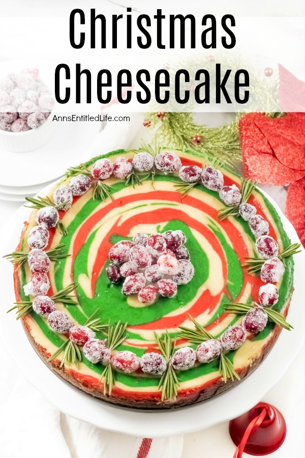 An overhead view of a round green and red swirled cheesecake topped with sugared cranberries and rosemary on a white serving dish. There is some holiday decor in the upper right, a bowl of sugared cranberries in the upper left.