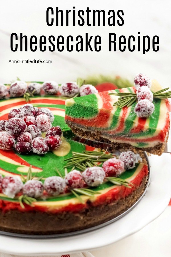 A round green and red swirled cheesecake topped with sugared cranberries and rosemary on a white serving dish. There is a piece being lifted from the whole cheesecake with a pie server.