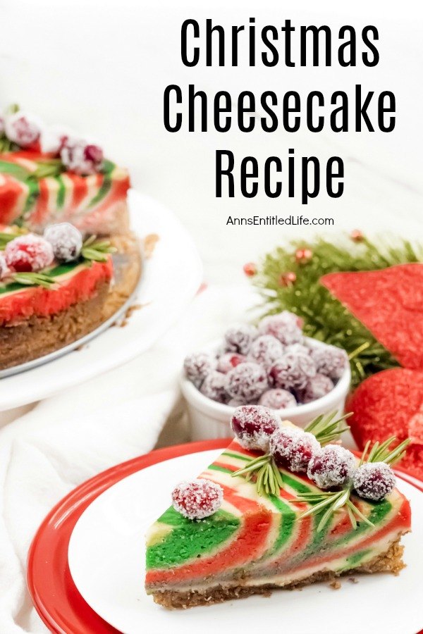 A piece of red and green Christmas cheesecake sits on a white plate stacked on top of a red plate, front and center. A white bowl of sugared cranberries is directly above it. The remaining cheesecake is on a white serving tray in the upper left.