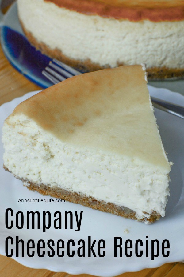 A slice of comany cheesecake site front and center on a white plate. The remaining cheesecake is underneath on a blue earthenwear dish in the background. This sits brown sideboard.