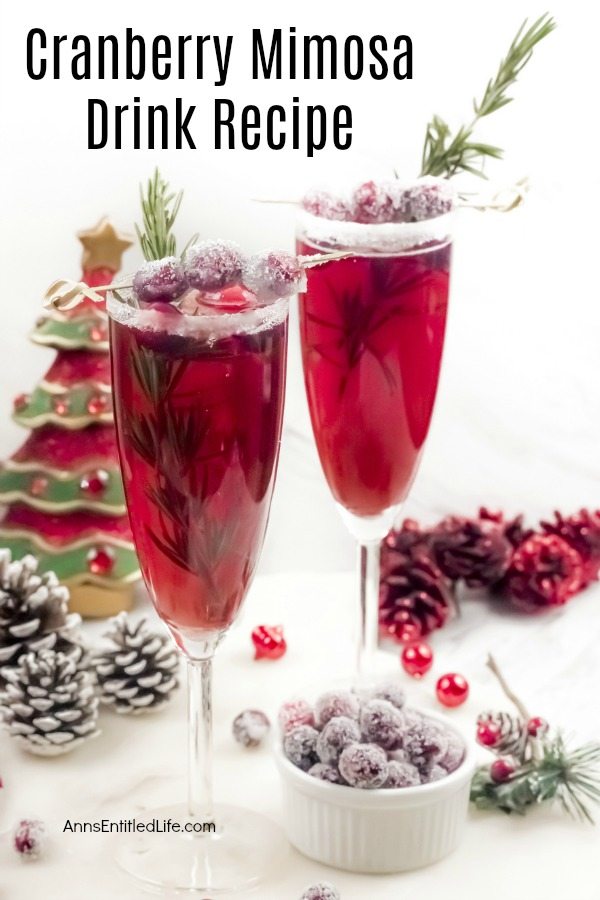 Two champagne flutes filled with cranberry mimosas, there is a white sugared rim, and garnished with rosemary inside the glass, and sugared cranberries on a stick on top. A white bowl filled with sugared cranberries sits at the bottom of the right flute, there is holiday decor on the white cupboard, and a faux Christmas tree in the back.