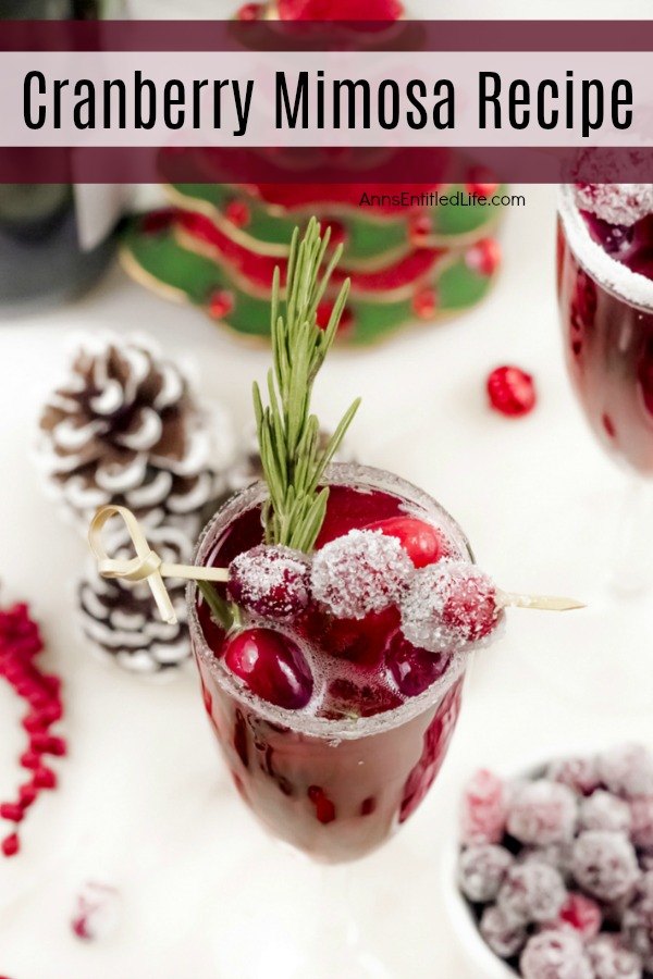 An overhead view of a champagne flute filled with cranberry mimosas, there is a white sugared rim, and garnished with rosemary inside the glass, and sugared cranberries on a stick on top. A white bowl filled with sugared cranberries sits at the bottom of the flute, there is holiday decor on the white cupboard, and a faux Christmas tree in the back.