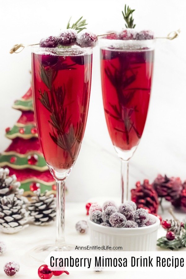 A close-up of two champagne flutes filled with cranberry mimosas, there is a white sugared rim, and garnished with rosemary inside the glass, and sugared cranberries on a stick on top. A white bowl filled with sugared cranberries sits at the bottom of the right flute, there is holiday decor on the white cupboard, and a faux Christmas tree in the back.