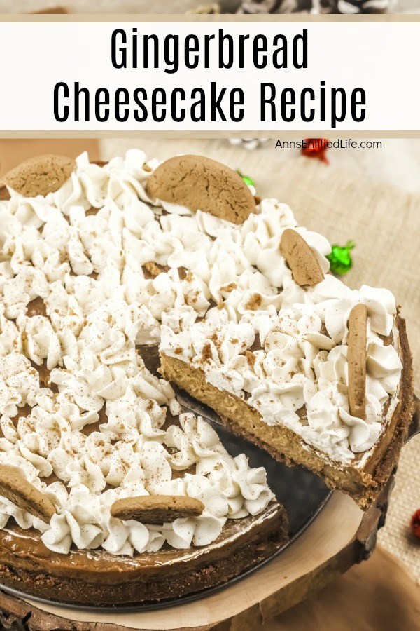 A whole gingerbread cheesecake sitting on a wooden serving tray on top of a brown countertop. A piece of the cheesecake is being lifted out with a pie server. The cheesecake is decorated with whipped cream and gingersnap cookies.