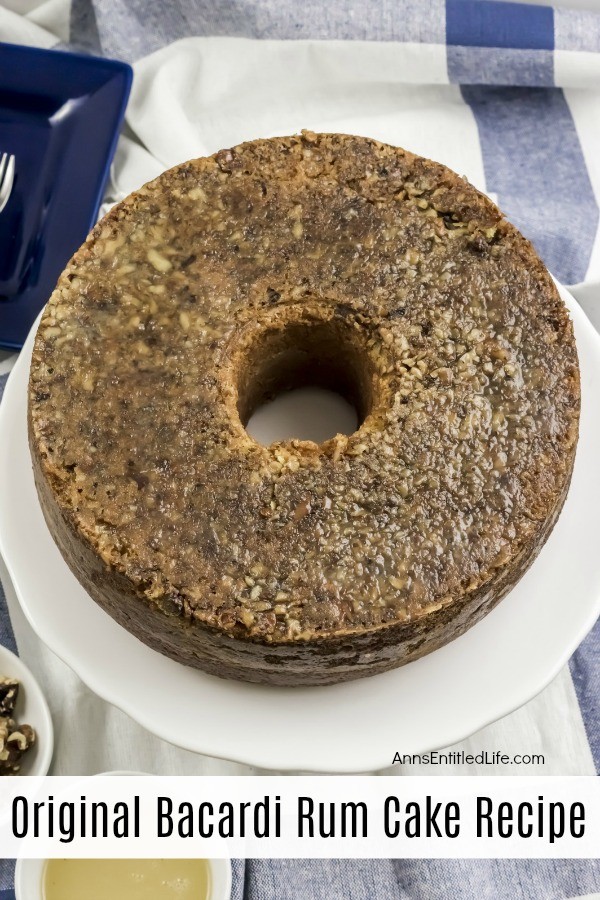 A whole Bacardi rum cake on a white cake server. There is a bowl of nuts in the center front. This is all on top of a white and blue bloth.