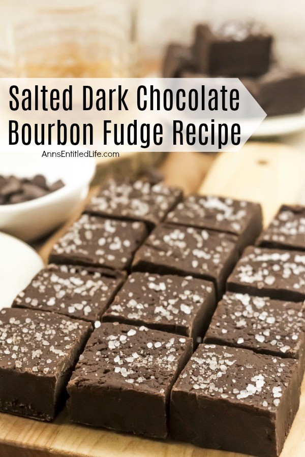  12 square pieces of salted dark chocolate bourbon fudge on a wooden cutting board which is set upon a gold napkin. There is a plate stacked with more fudge in the background.