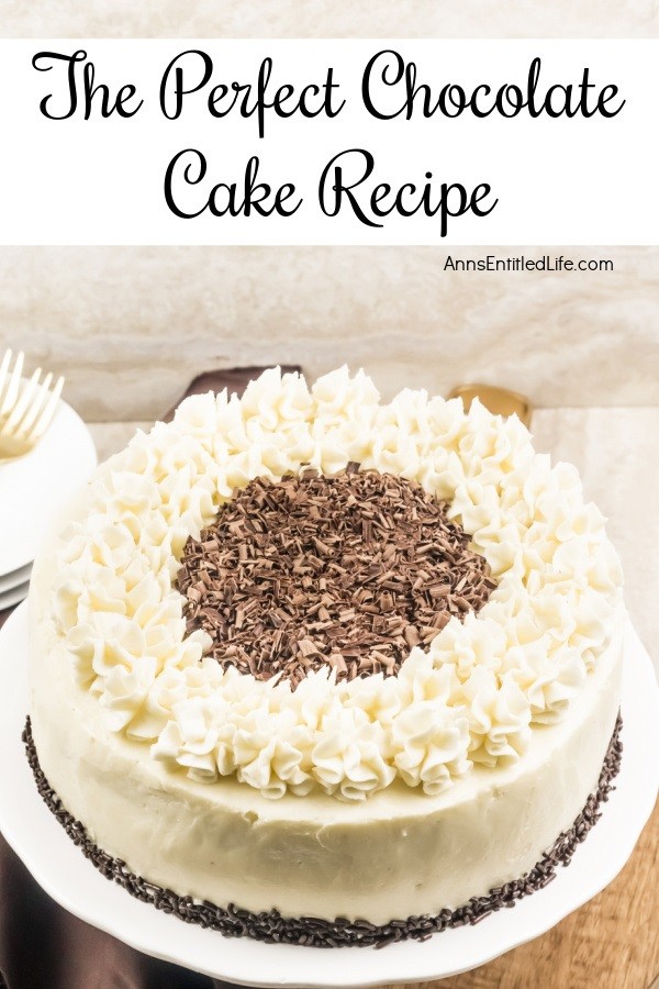 A side view of a white frosted cake trimmed with ruffle mounds, and chocolate shavings in the middle, on a white cake plate. There is a small stack of white plates and some gold silverware in the upper left.