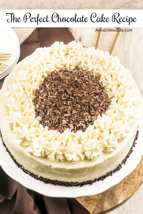 An overhead view of a white frosted cake trimmed with ruffle mounds, and chocolate shavings in the middle, on a white cake plate. There is a small stack of white-colored plates and some gold silverware in the upper left.