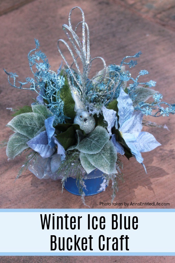 a blue bucket with ice blue and silver floral, and a silver bird, arrangement placed on the concrete outside.