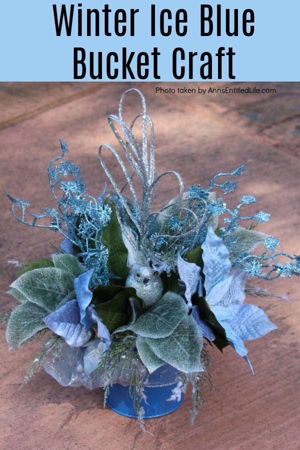 a blue bucket with ice blue and silver floral, and a silver bird, arrangement placed on the concrete outside.