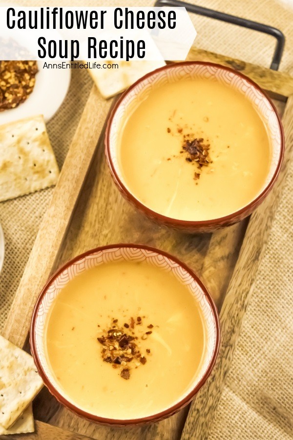 Two brown soup bowls on a brown wooden serving tray are filled with cauliflower cheese soup. There are crackers to the left, and a bowl of pepper flakes in a white bowl to the upper left.