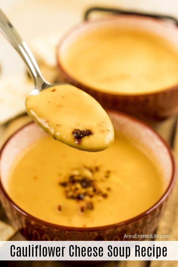 Two brown soup bowls on a brown wooden serving tray are filled with cauliflower cheese soup. A spoon full of soup is being lifted from the first bowl. There are crackers to the left, and a bowl of pepper flakes in a white bowl to the upper left.