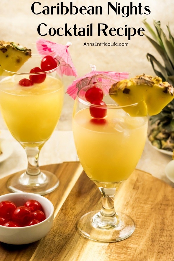 Front view of two Caribbean Nights cocktails garnished with pineapple and cherries and cocktail umbrellas a small white bowl is filled with pineapple in the upper right, and a small right bowl is filled with cherries in the lower left. This is on a butcher block tray.