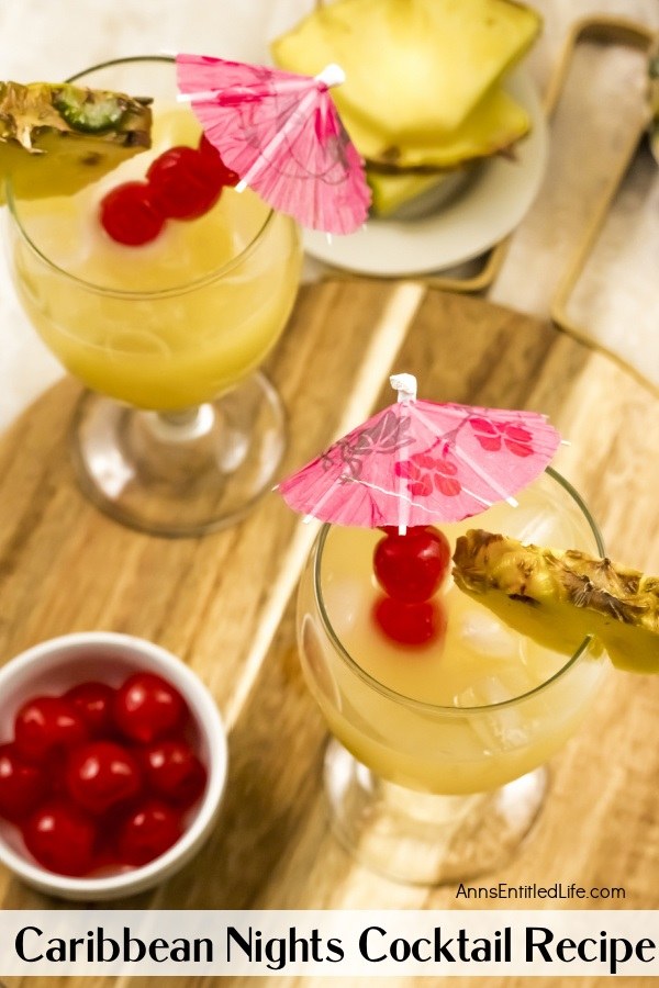 overhead of two Caribbean Nights cocktails garnished with pineapple and cherries and cocktail umbrellas a small white bowl is filled with pineapple in the upper right, and a small right bowl is filled with cherries in the lower left. This is on a butcher block tray.