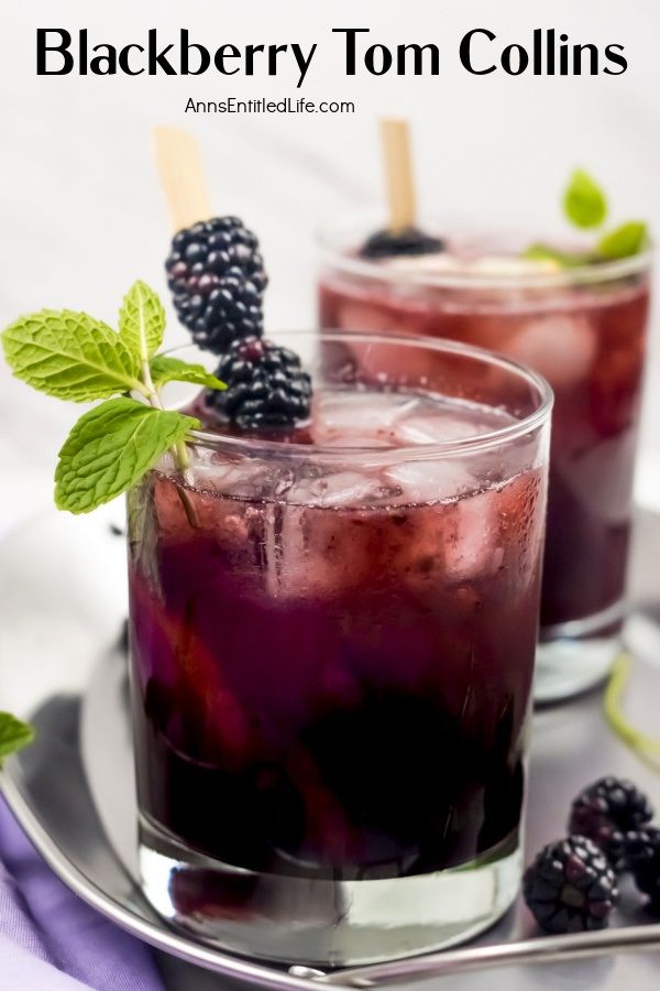 A blackberry Tom Collins garnished with blackberries, mint, and lemons againsta white background. A second blackberry Tom Collins is in the background.