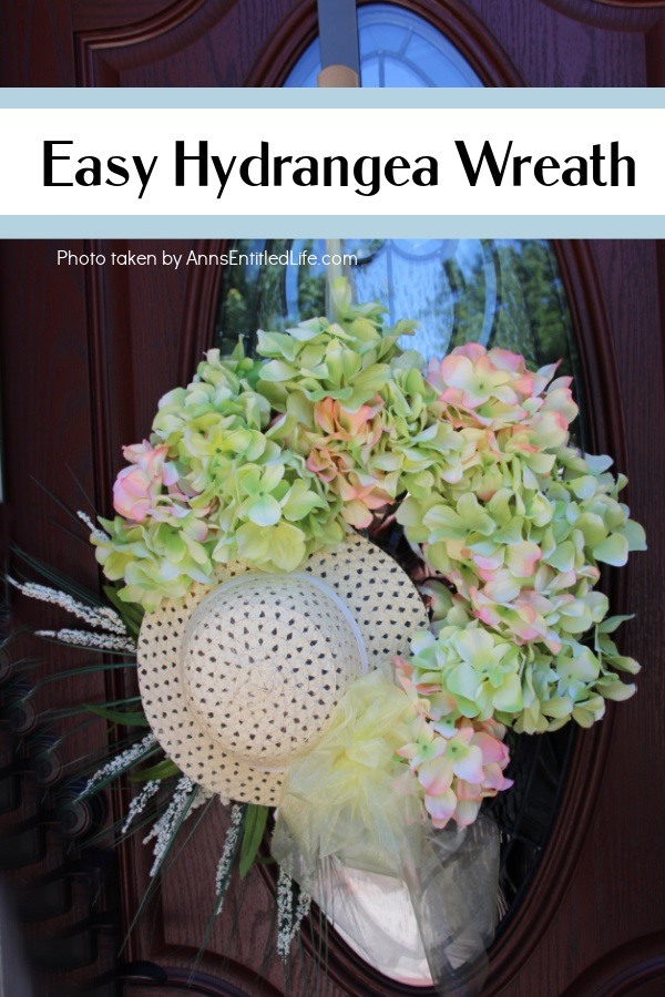 A left side photo of a Hydrangea Wreath made with a hat, hanging off a door hanger against a brown door with a glass window.