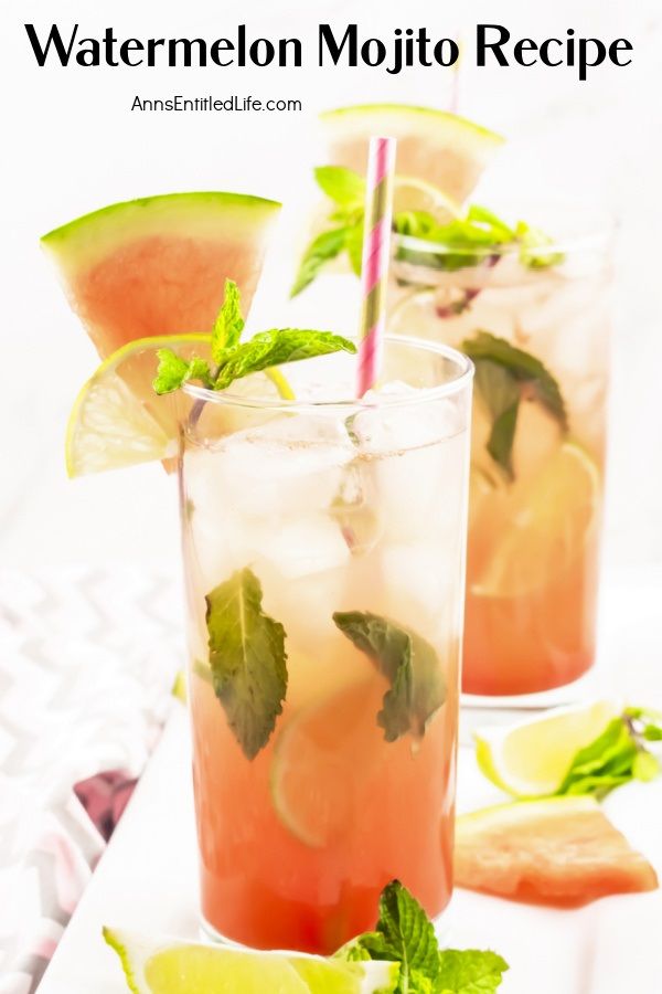 Two watermelon mojitos, one cocktail is front and center, on a white tray surrounded by cut watermelon, lime slices, and min springs. This sits on a pink and grey chevron napkin.