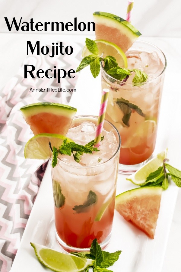 Two watermelon mojitos on a white tray surrounded by cut watermelon, lime slices, and min springs. This sits on a pink and grey chevron napkin.