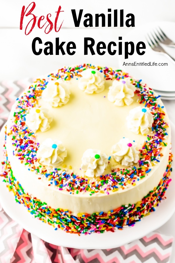 A whole vanilla cake frosted with buttercream frosting, trimmed with ruffle mounds, and decorated with colored sprinkles, on a white cake plate. There is a small stack of white-colored plates and some silverware in the upper right. All this sits on a pink and grey chevron napkin.