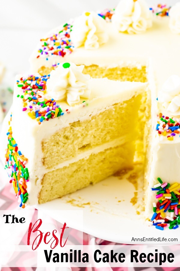 A piece of vanilla cake frosted with buttercream frosting, trimmed with ruffle mounds, and decorated with colored sprinkles is being lifted from the remainder of the cake which sits on a white cake plate. There is a small stack of white-colored plates and some silverware in the upper right. All this sits on a pink and grey chevron napkin.