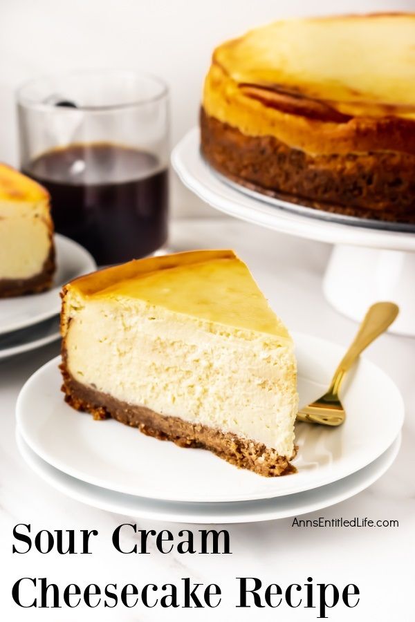 Close-up of a piece of sour cream cheesecake on a white plate, gold fork on the plate. The remaining cheesecake is on a white cakedish in the background, a cup of coffee to the left.