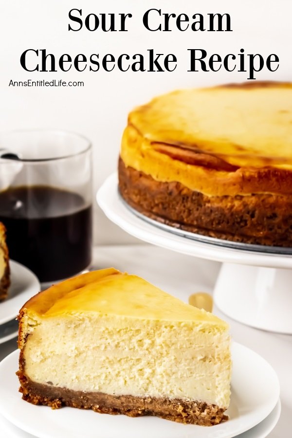 Close-up of a piece of sour cream cheesecake on a white plate, gold fork on the plate. The remaining cheesecake is on a white cakedish in the background, a cup of coffee to the left.