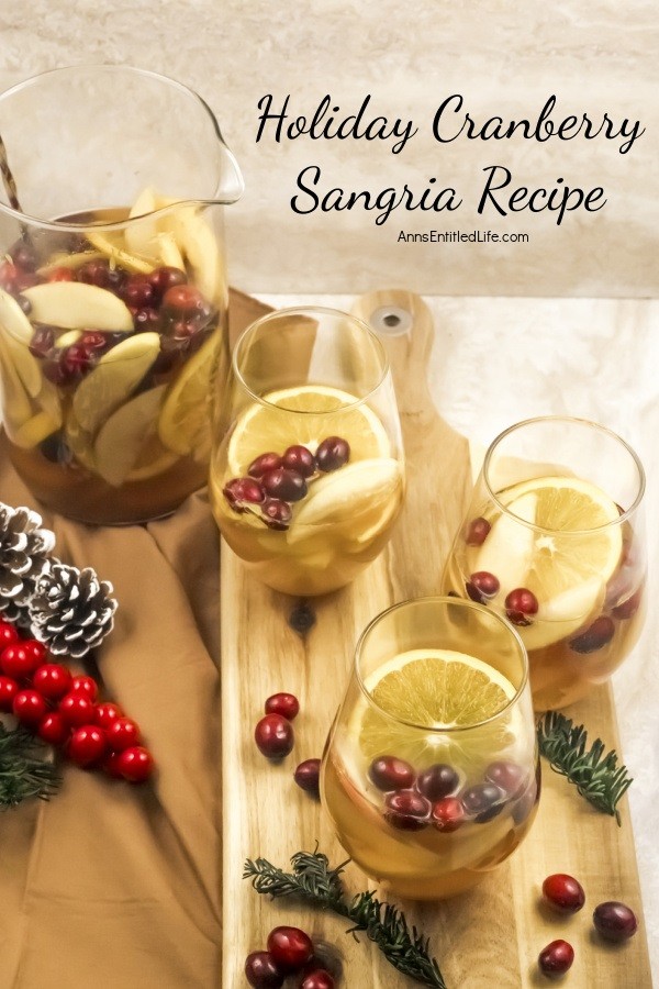 Holiday Cranberry Sangria Recipe Three glasses half-filled with holiday cranberry sangria and garnished with orange slices and cranberries sitting on a wooden board. The remaining sangria is in a clear pitcher in the upper left. There are cranberry and rosemary garnishes sitting on the board as decorations