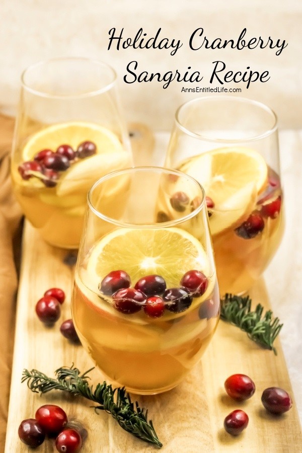 Three glasses half-filled with holiday cranberry sangria and garnished with orange slices and cranberries sitting on a wooden board. There are cranberry and rosemary garnishes sitting on the board as decorations