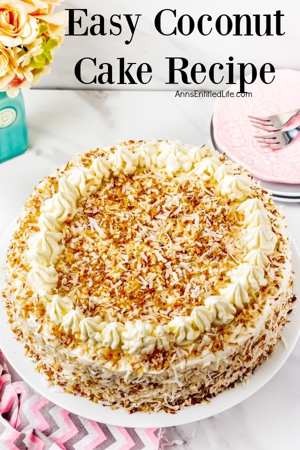 A whole coconut cake trimmed with ruffle mounds and decorated with toasted coconut, on a white cake plate. There is a small stack of plates and some silverware in the upper right. All this sits on a pink and grey chevron napkin.