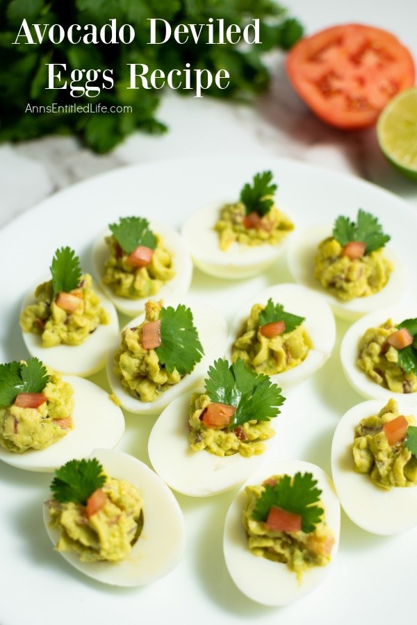 a dozen avocado deviled eggs on a white plate, half a tomato and half a lime in the upper right