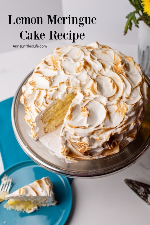 A whole lemon meringue cake on a silver platter with one piece removed. That piece is on a blue plate, lower left with a fork sitting beside it.
