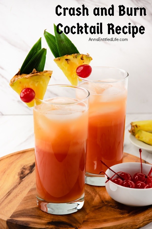 Two crash and burn cocktails in tall glasses garnished with pineapple and cherries on a wooden board. There is a white bowl of cherries on the right, a white bowl of pineapple in the upper right.